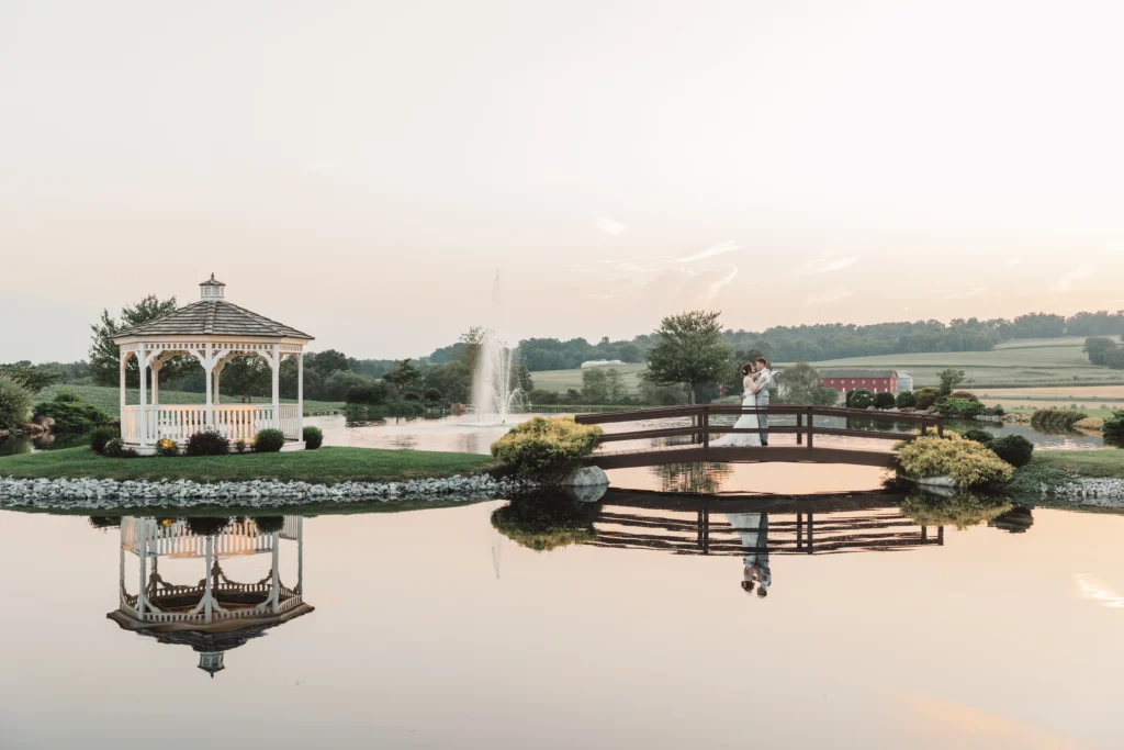 bride and groom embracing on bridge above pond with gazebo on the left
